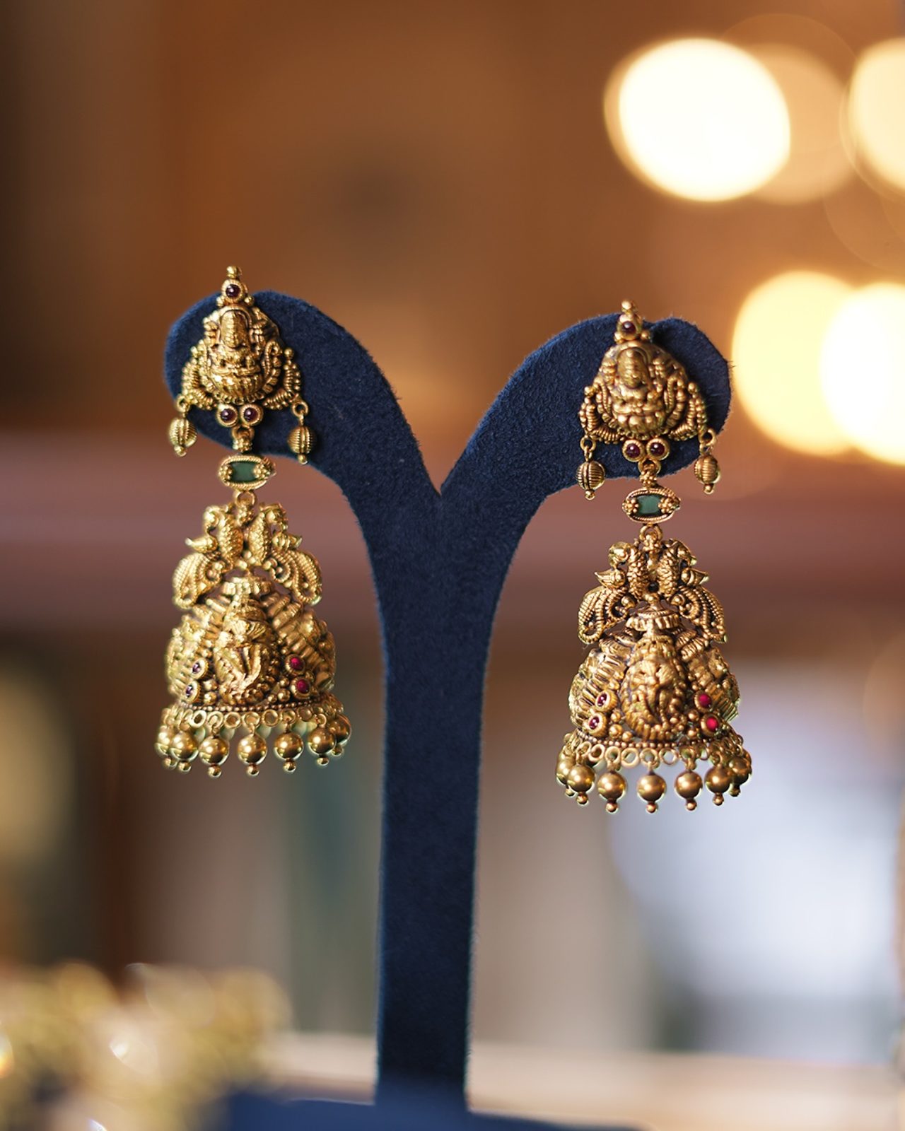 Lakshmi jhumka temple earrings in antique gold against warm showroom bokeh